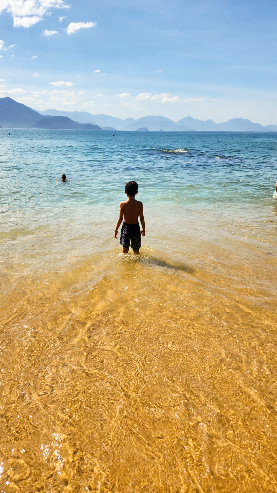 Thales olhando o mar com os pés na parte rasa da praia.
